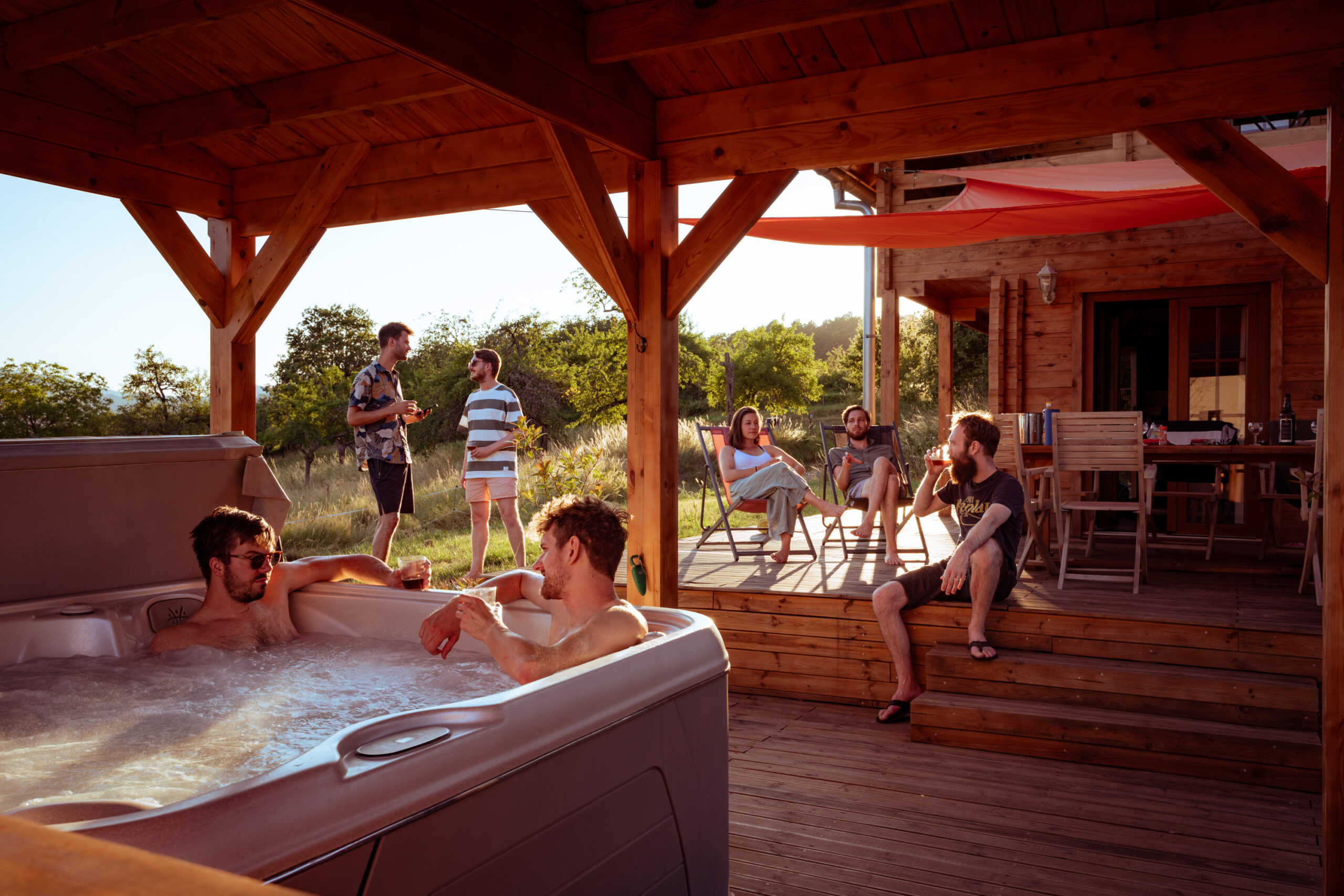 Een vriendengroep geniet van de avondzon bij hun houten chalet met jaccuzzi, midden in de natuur. De sfeer is warm, ontspannen en gezellig.